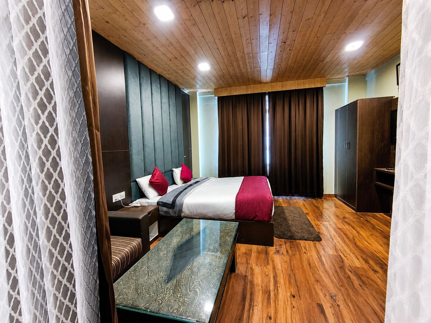 A modern hotel room with a wooden ceiling, a bed with red and white bedding, a glass table, a seating area, and a wardrobe.