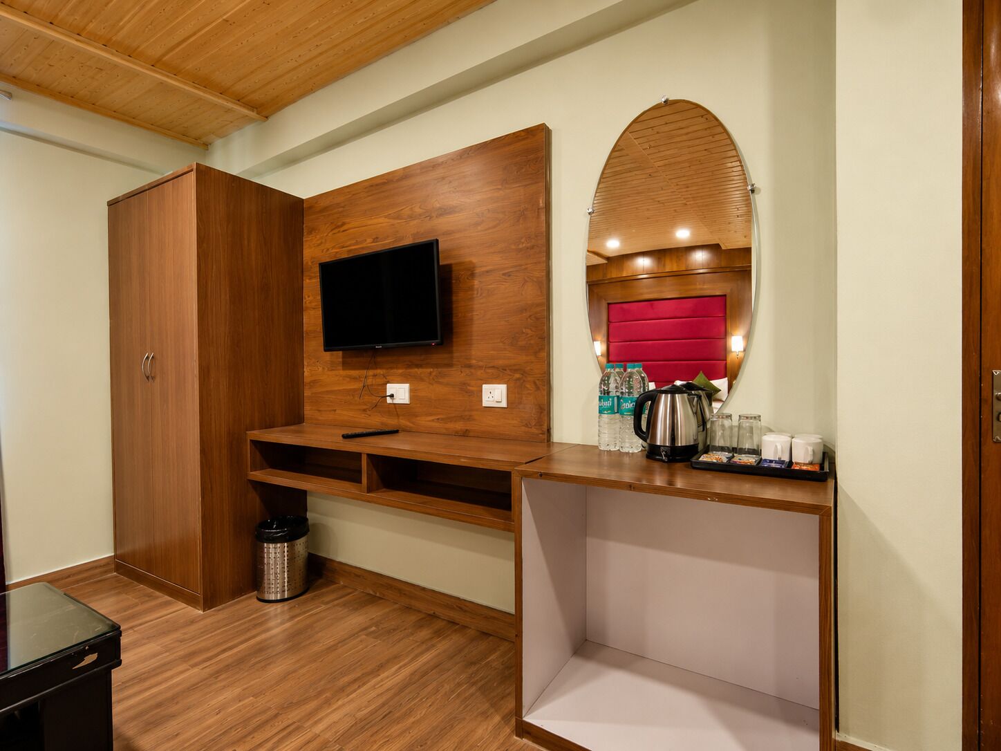 A hotel room with a wooden wardrobe, a mounted TV, a red cushioned headboard, a coffee station, and a trash bin.