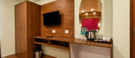 A hotel room with a wooden wardrobe, a mounted TV, a red cushioned headboard, a coffee station, and a trash bin.