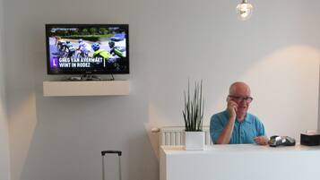 A man sitting at a reception desk with a television mounted above displaying a cycling event.