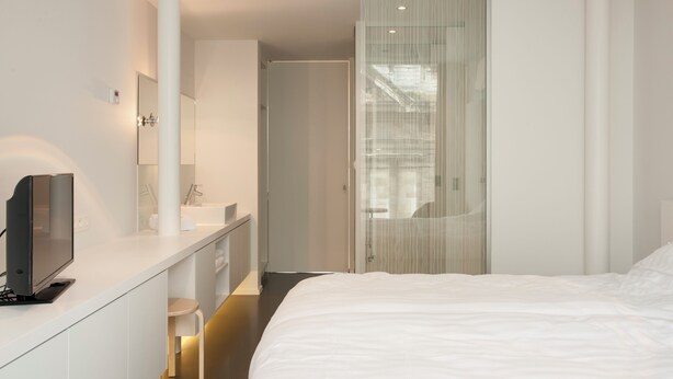 Une chambre d’hôtel moderne avec un grand lit, un téléviseur posé sur une console blanche, une salle de bain avec un miroir et un lavabo, et une porte coulissante donnant sur une autre pièce.