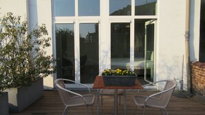 Une terrasse avec une table et deux chaises, un pot de fleurs et une vue sur un immeuble à travers de grandes fenêtres.