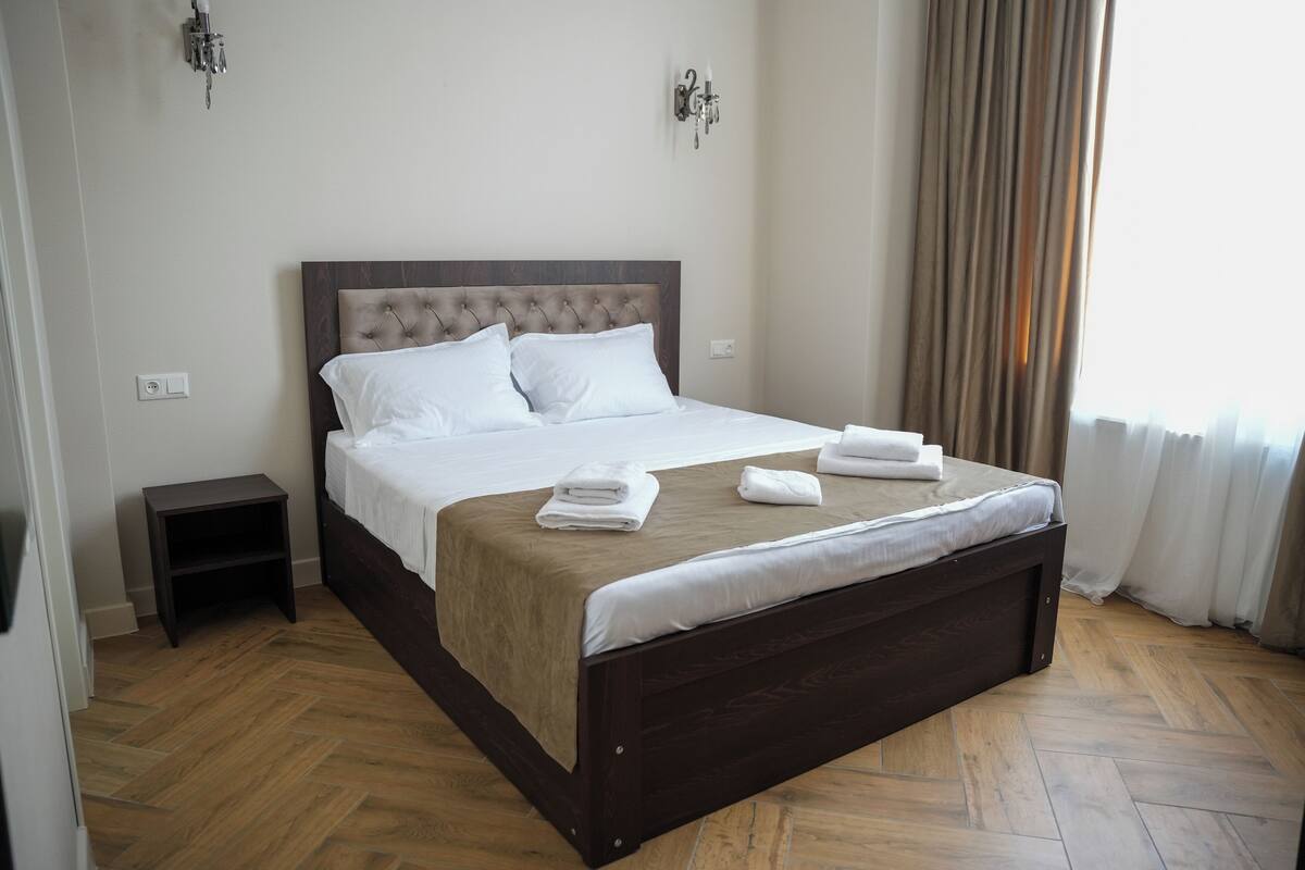 Een opgemaakt bed met witte lakens en handdoeken, een donker houten hoofdbord en een klein nachtkastje.