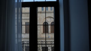 Blick durch ein Fenster mit Gitterrost, zeigt eine Gebäudefassade mit Rundbogenfenstern und einem Balkon mit schmiedeeisernem Geländer.