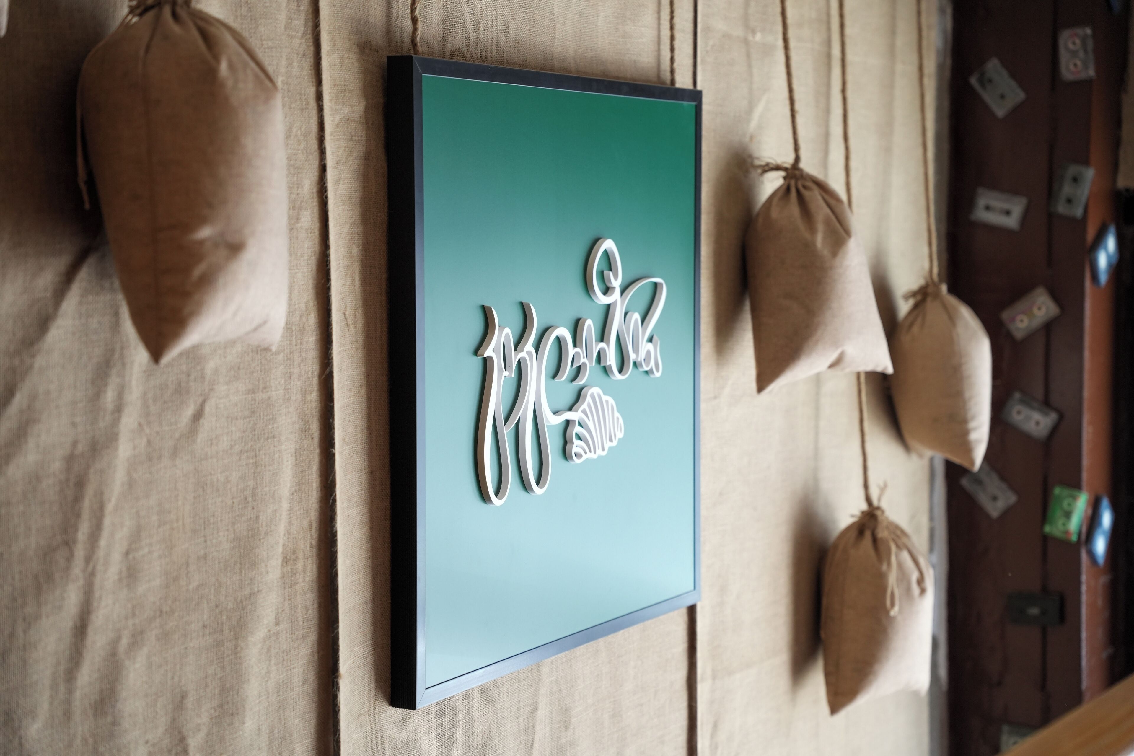 A framed artwork with the word "welcome" in stylized script, hanging on a wall with a beige fabric backdrop.