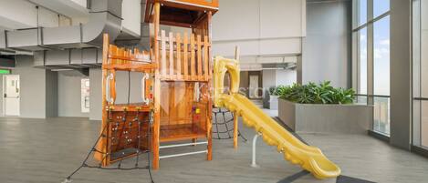 A play area with a wooden climbing structure and a yellow slide in a spacious indoor setting.