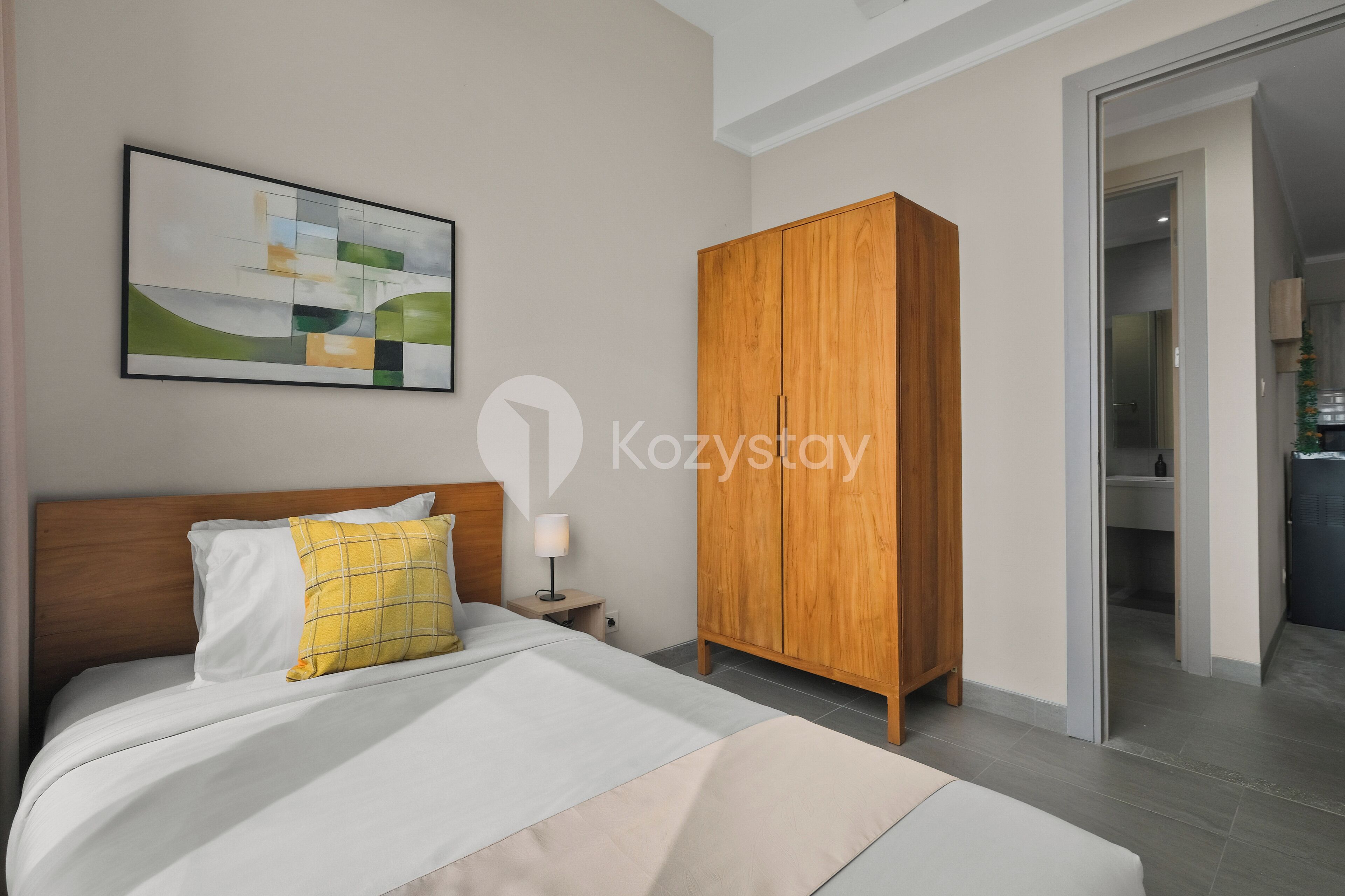 A hotel room with a bed, a wooden wardrobe, a nightstand with a lamp, and a painting on the wall.