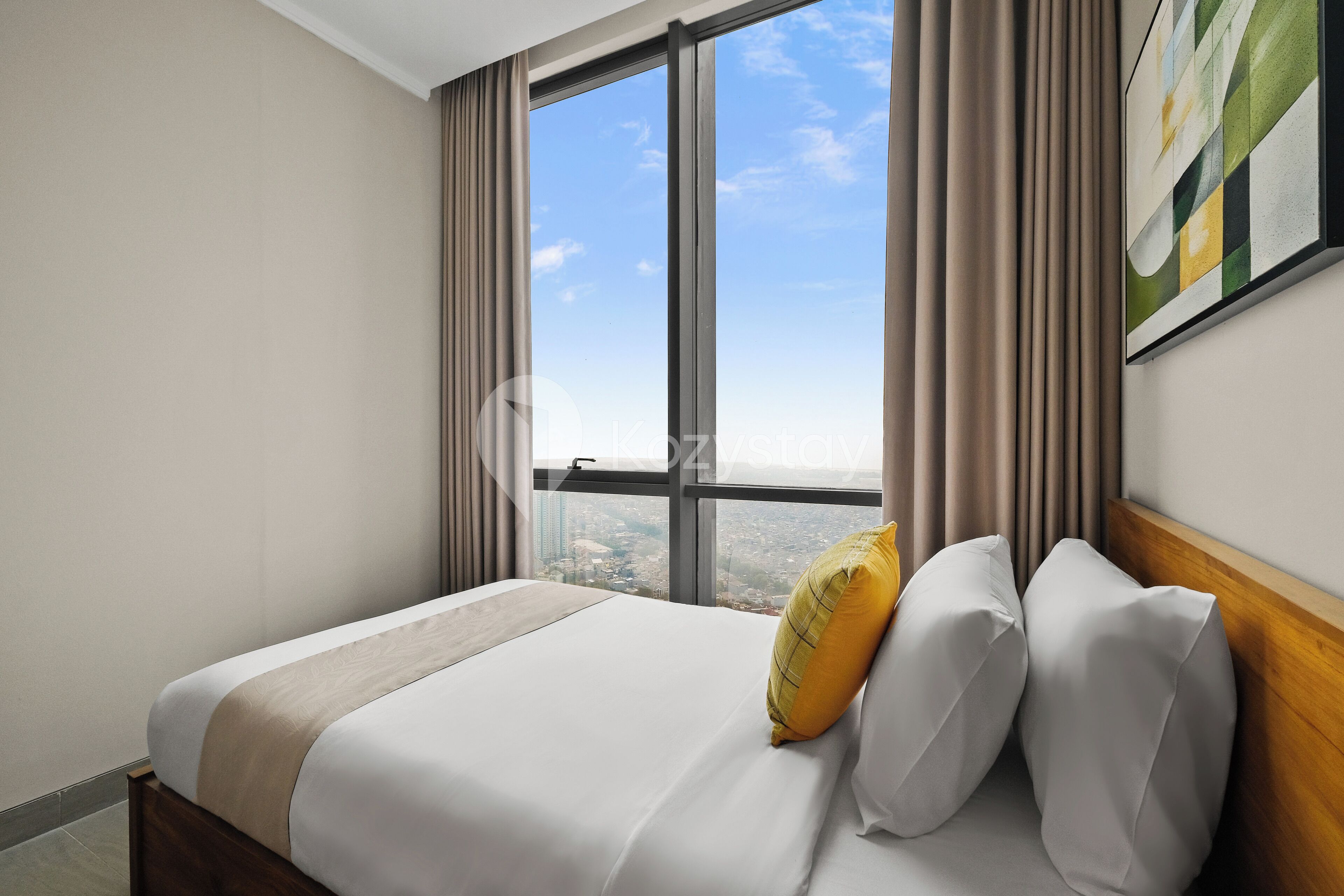 Ein Hotelzimmer mit großem Fenster, einem Bett mit weißer Bettwäsche und einem gelben Kopfkissen, sowie einem Gemälde an der Wand.