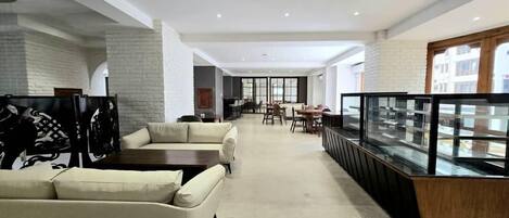 A modern hotel lobby with a seating area, a glass display case, and a dining area in the background.