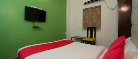 A room with a bed, a television mounted on the wall, a window with a patterned blind, and an air conditioning unit.