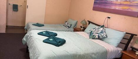 A hotel room with two beds, each with teal bedding and pillows, a nightstand with a lamp, and a painting of a sunset on the wall.