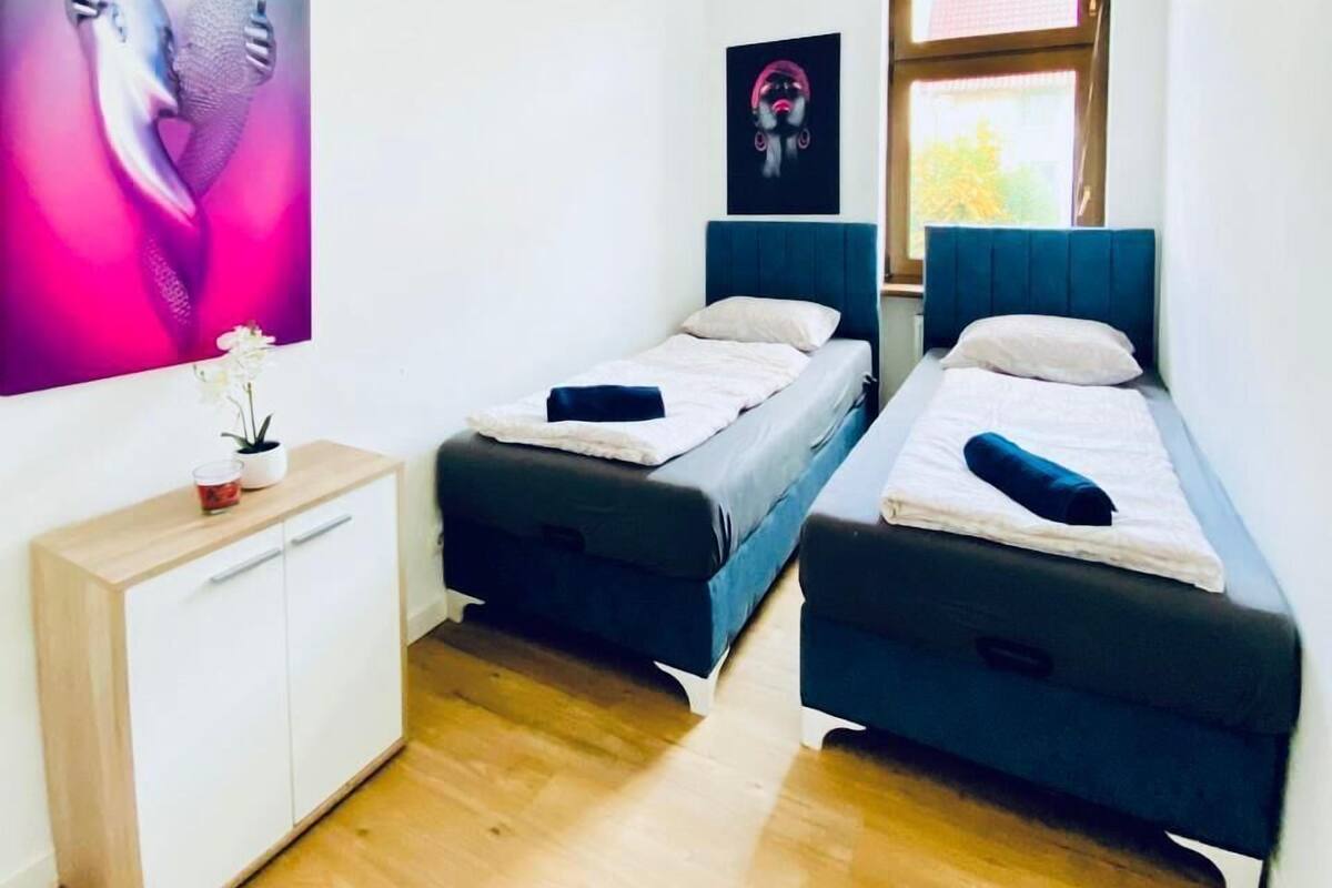 Une chambre moderne avec deux lits simples, une commode en bois et un grand tableau abstrait accroché au mur.