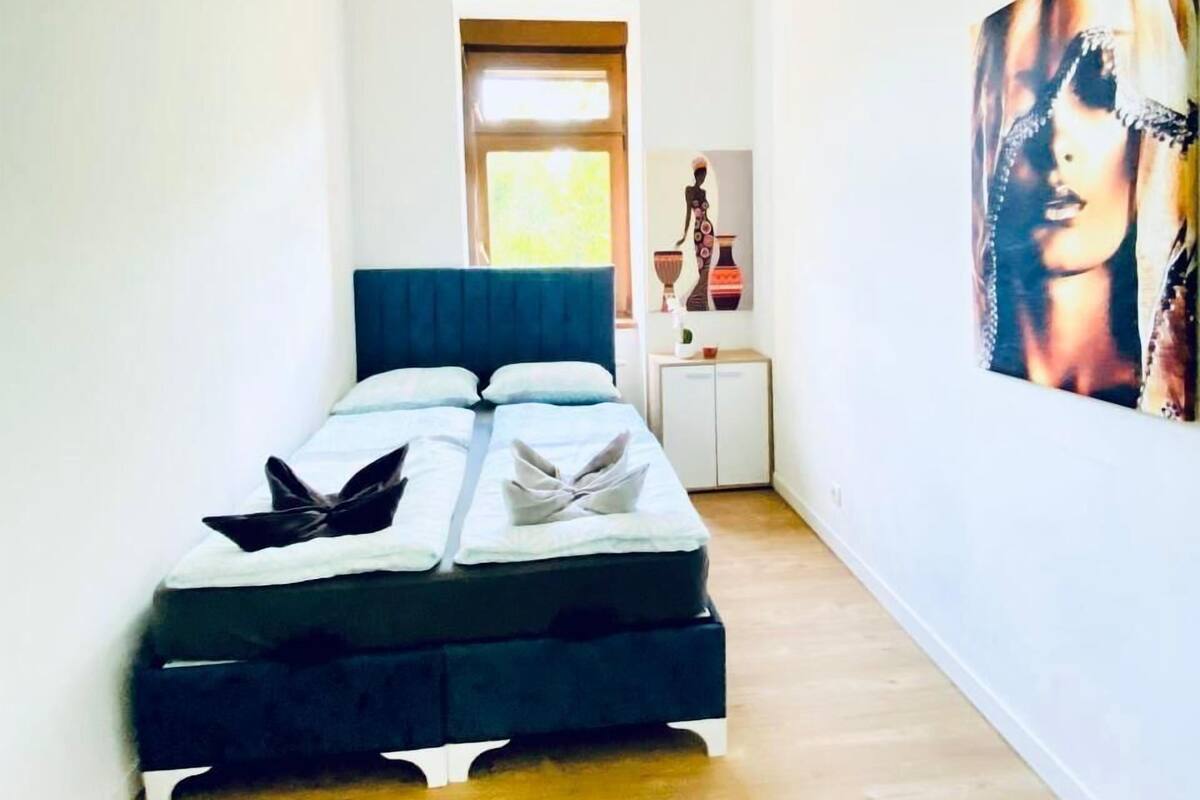 Une pièce étroite avec un lit, une petite table sur laquelle se trouve un vase, et un tableau accroché au mur.