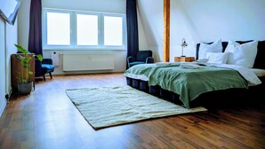 A modern bedroom with a large window, a bed with white linens, a blue chair, and a wooden floor.