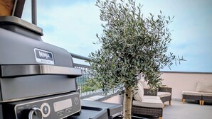 A NINJA woodfire grill on an outdoor patio with a potted olive tree and wicker furniture.