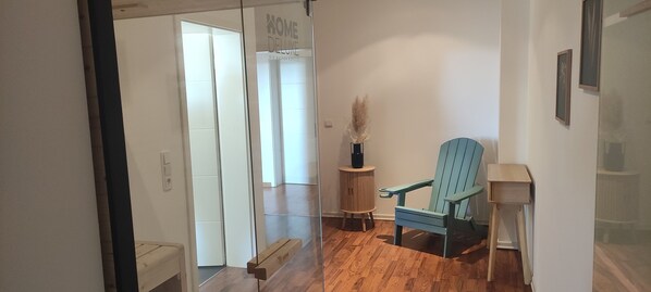 Um corredor com piso de madeira, uma cadeira azul, uma pequena mesa com um vaso e uma porta com a inscrição