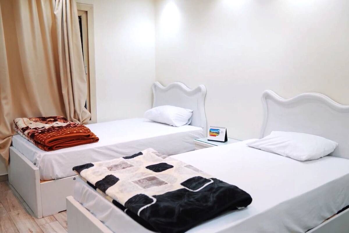 A hotel room with two single beds, each with a white headboard and a white mattress. There is a small bedside table with a calendar on it.