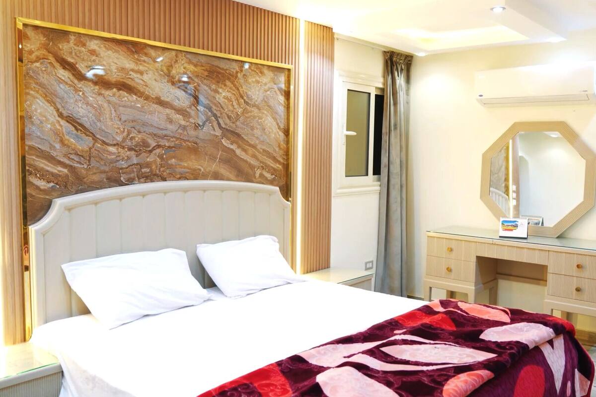 A hotel room with a marble headboard, a bed with white linens, a wooden dresser, and a large mirror.