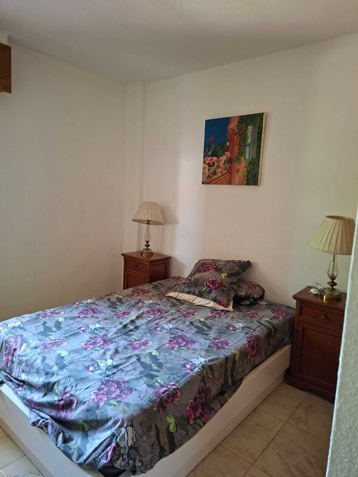 A bedroom with a bed, two nightstands, a wall-mounted lamp, and a painting.