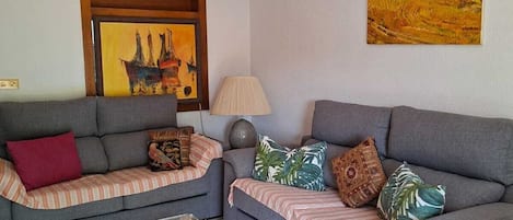 Una sala de estar con dos sofás grises, una mesa de centro de vidrio transparente, un ventilador de techo, una lámpara y dos cuadros.