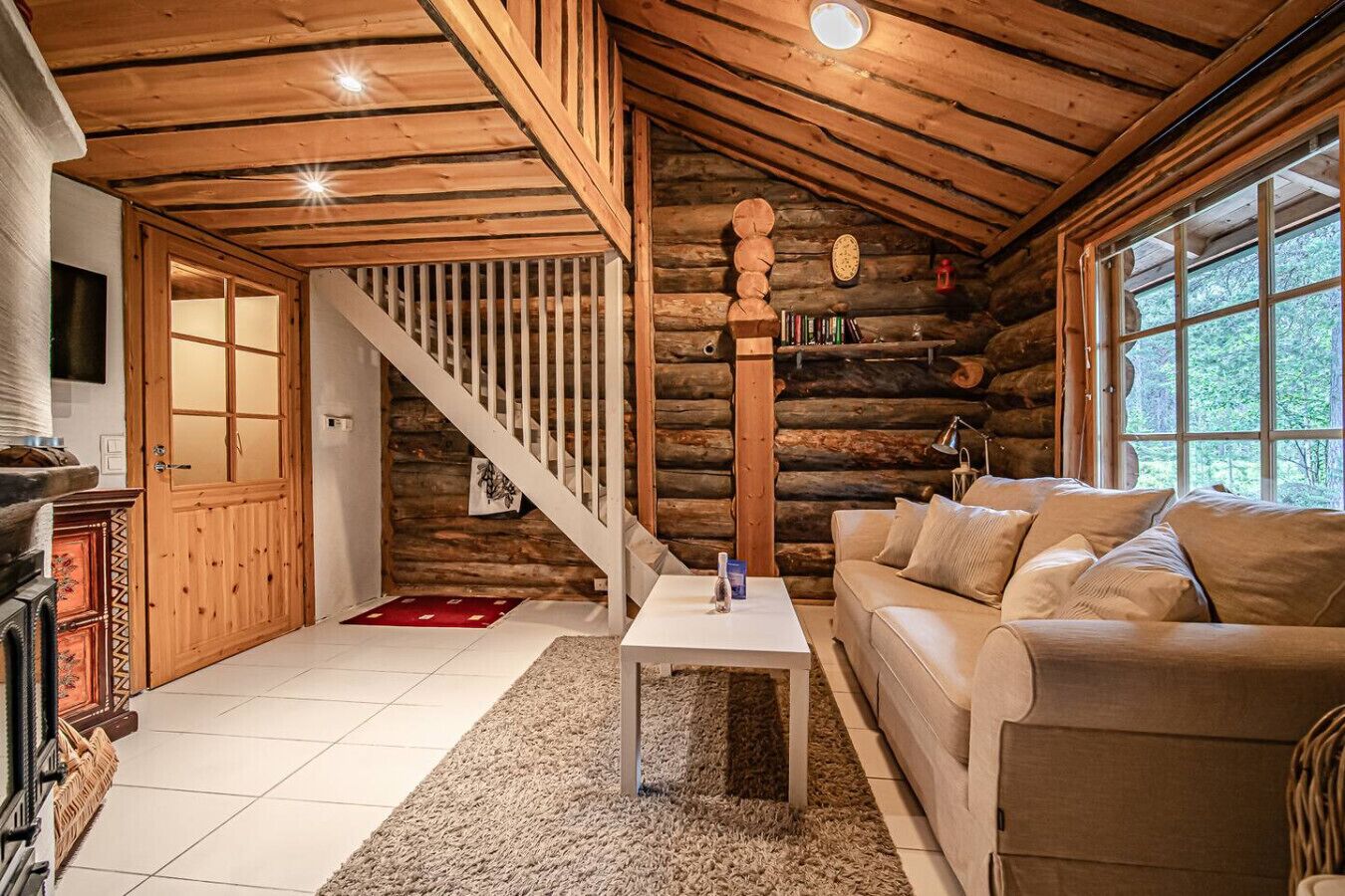 Interior de una cabaña de troncos con escalera, sofá, mesa y chimenea.