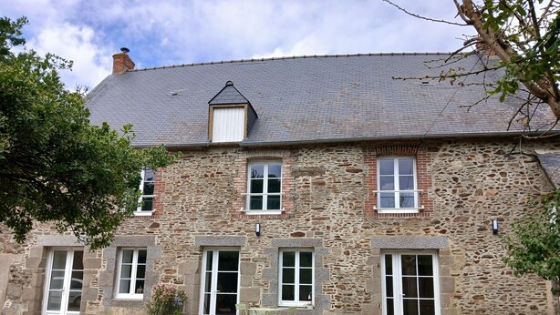 A stone building with a sloped roof, multiple windows, and a small outdoor seating area with chairs. - Maison breronne (Meillac)