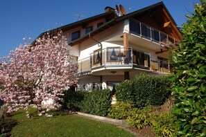 Un bâtiment à deux étages doté d’un balcon, entouré de jardins bien entretenus et d’un arbre en fleurs.