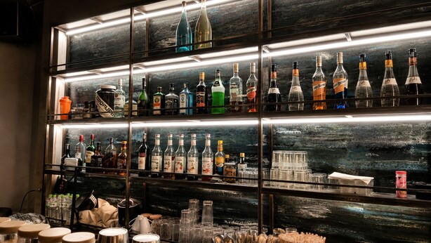 A bar with multiple shelves of liquor bottles, glasses, and a backdrop of a cityscape.