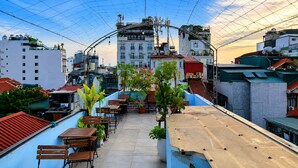 A rooftop terrace with wooden tables and chairs, potted plants, and a view of surrounding buildings.