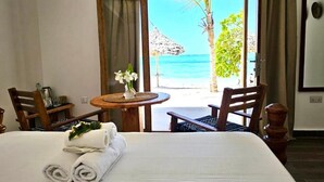 A hotel room with a bed, two chairs, a small table with a vase of flowers, and a view of the beach and ocean.