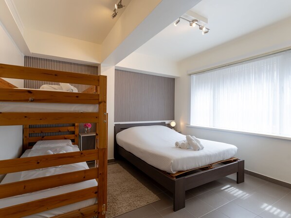 A modern bedroom with a bunk bed, a single bed, a bedside table with a lamp, and a large window with blinds.