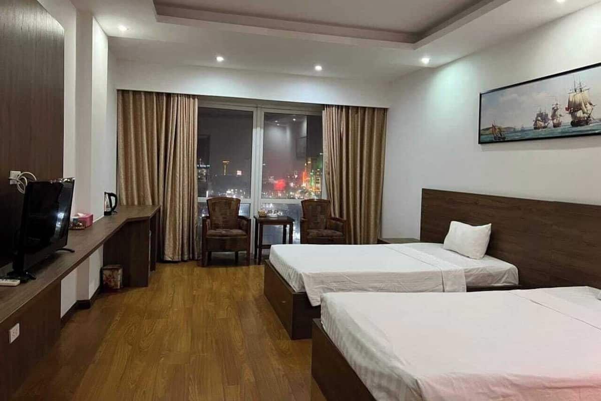 Quarto de hotel com duas camas, televisão, escrivaninha e vista da cidade à noite.