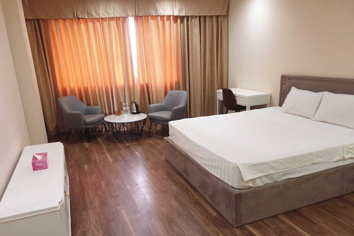 Quarto de hotel com uma cama grande, duas poltronas, uma mesinha redonda com uma chaleira e uma escrivaninha com cadeira.