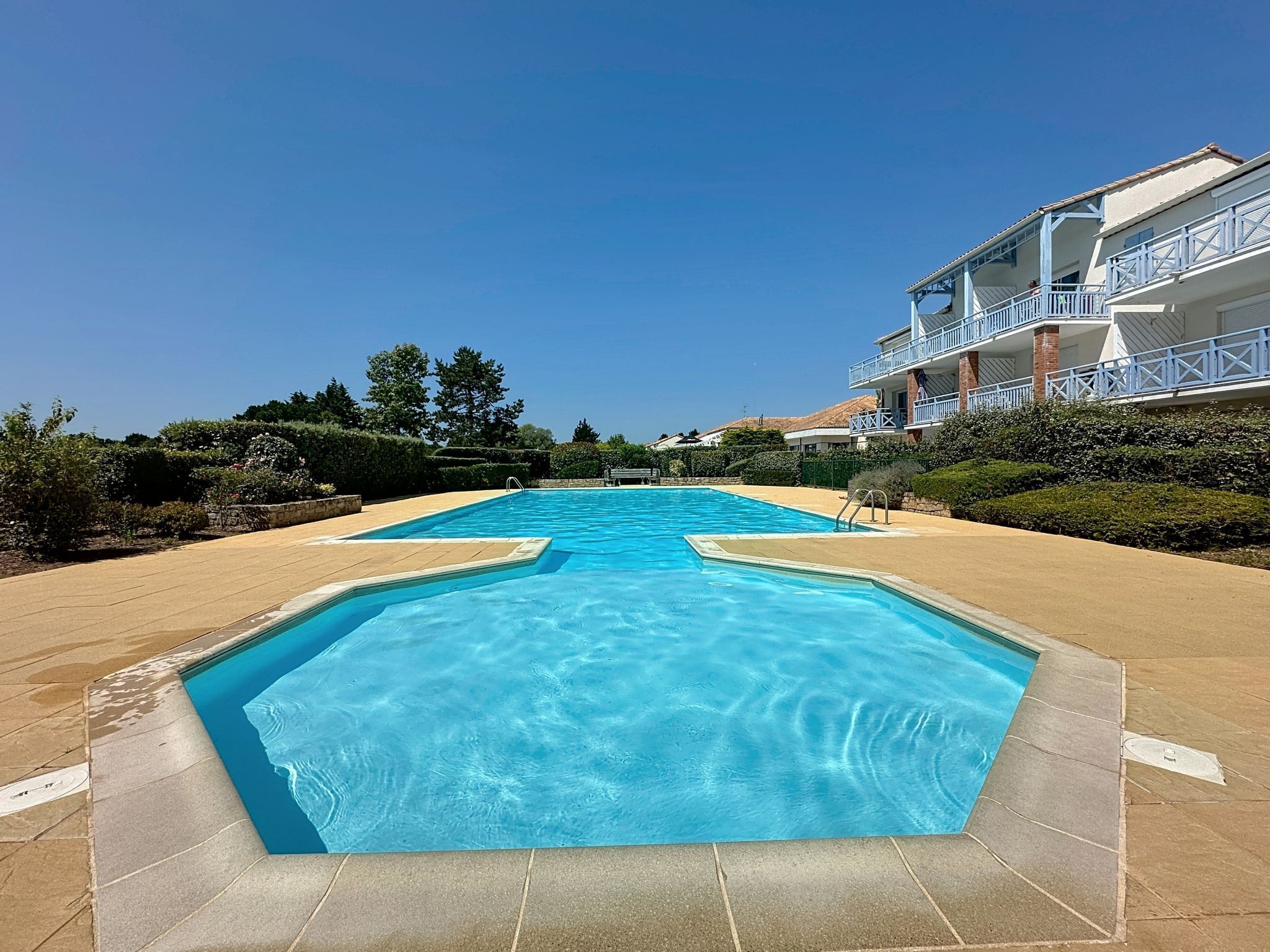 A swimming pool with a clear blue water surface, surrounded by a paved area and a building with balconies.