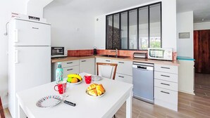 A modern kitchen with white cabinets, a built-in oven, microwave, and dishwasher. There is a dining table with plates, a knife, and a fruit bowl.