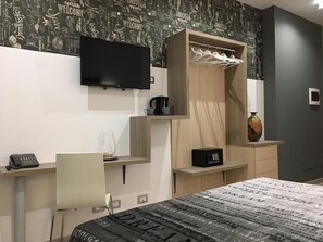 Ein modernes Hotelzimmer mit Bett, Schreibtisch, Stuhl, Wandfernseher, Kleiderschrank mit Kleidung und einem kleinen Tisch mit Kaffeemaschine.