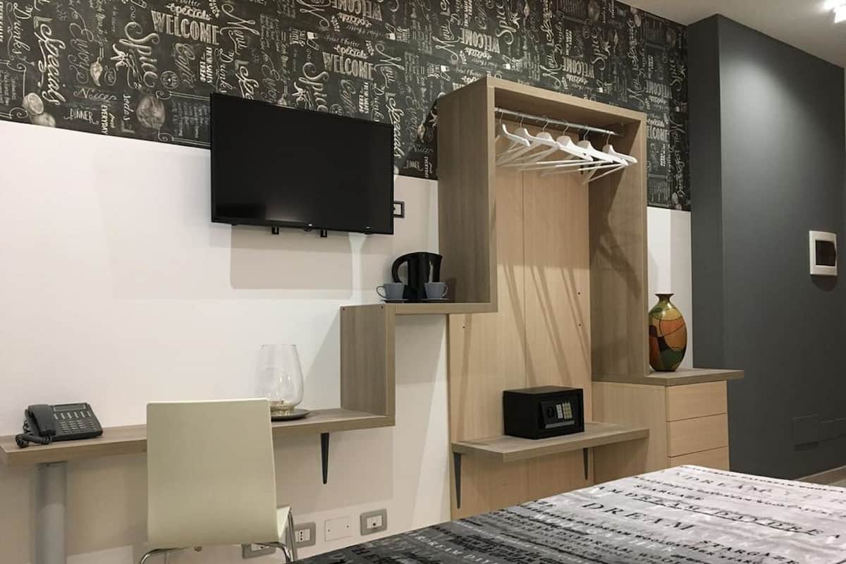 Une chambre d’hôtel moderne dotée d’un téléviseur à écran plat fixé au mur, d’une armoire en bois où sont suspendus des vêtements, d’un bureau avec un téléphone et d’un lit recouvert d’une couvre-lit à motifs.