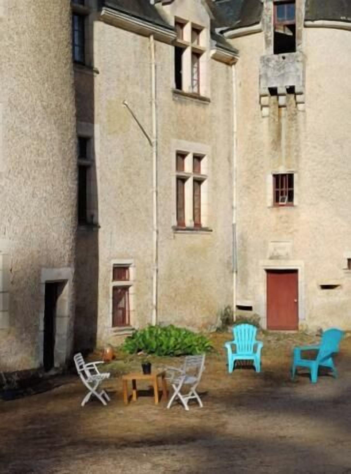 A courtyard with a stone building, a red door, and outdoor furniture including two blue chairs and a table.