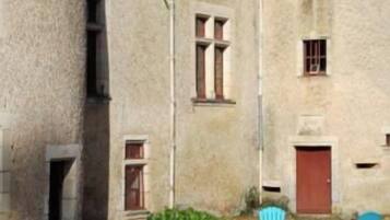 Une cour avec un bâtiment en pierre, une porte rouge et du mobilier d’extérieur comprenant deux chaises bleues et une table.