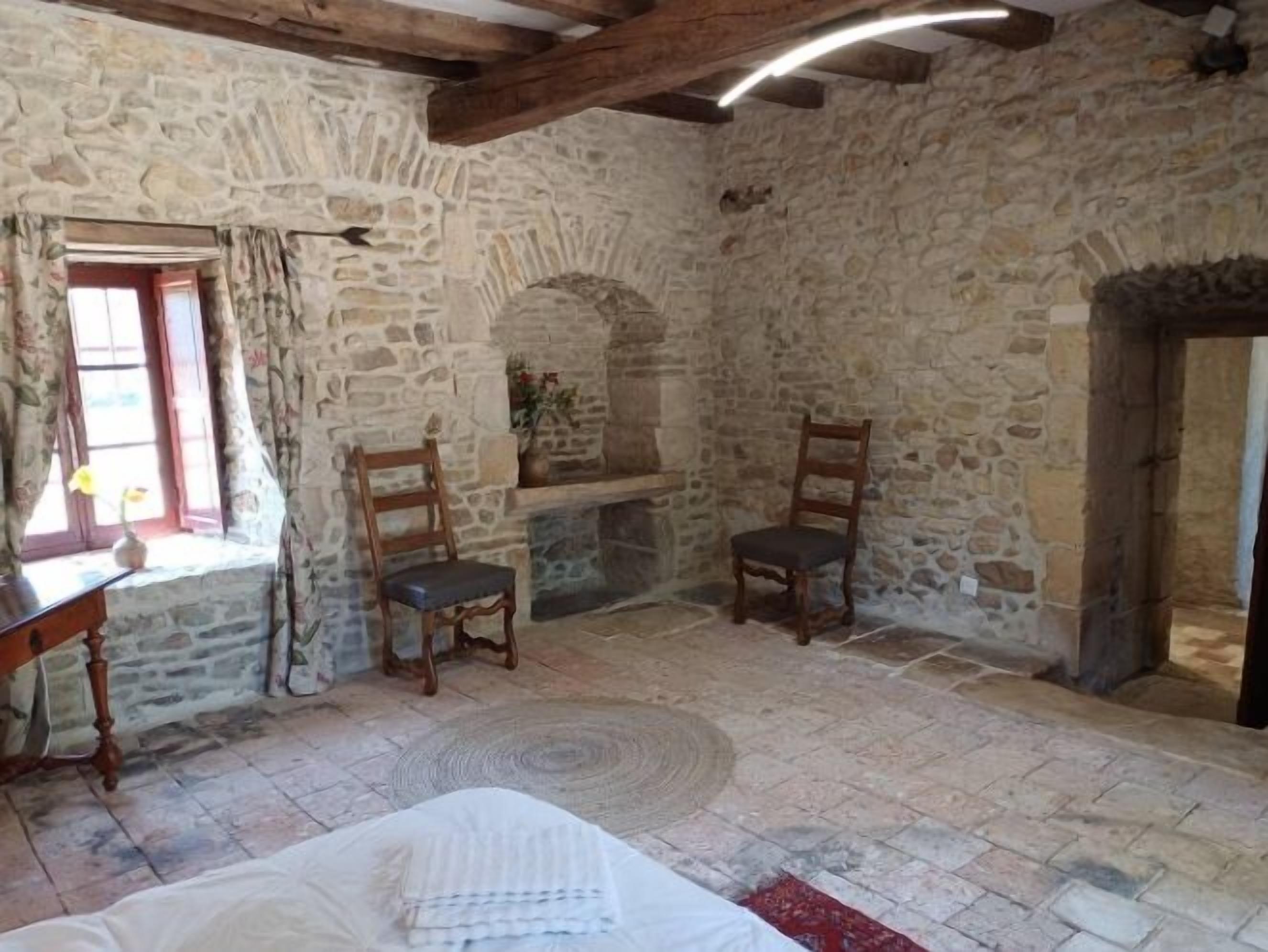 A stone-walled room with two wooden chairs, a fireplace, and a bed with a white cover.