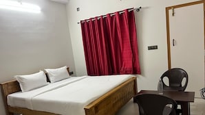 A bedroom with a wooden bed, white bedding, a red curtain, a black chair, and a small table with a bottle.
