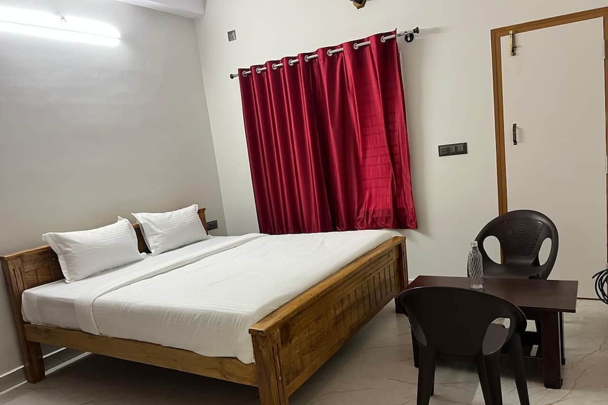 A bedroom with a wooden bed, white bedding, a red curtain, a black chair, and a small table with a bottle.