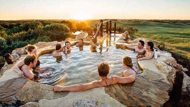 Un groupe de personnes se détend dans un cadre naturel, au bord d’une source d’eau chaude, avec un coucher de soleil en arrière-plan.