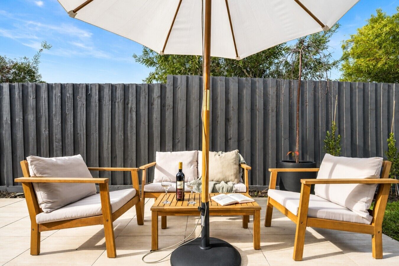 An outdoor patio area with wooden furniture, a table with a bottle and glasses, and a large umbrella.