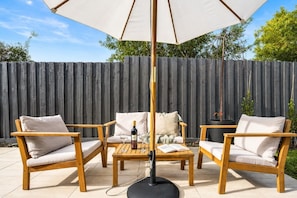 Un espacio de patio al aire libre con mobiliario de madera, una mesa con una botella y vasos, y un paraguas grande.