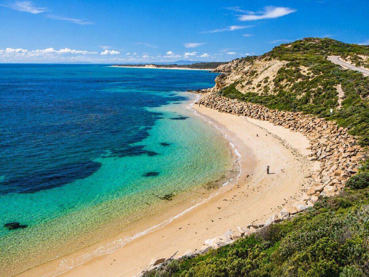 Eine Küstenlandschaft mit klarem Strand, felsiger Uferlinie und einer kurvenreichen Straße.