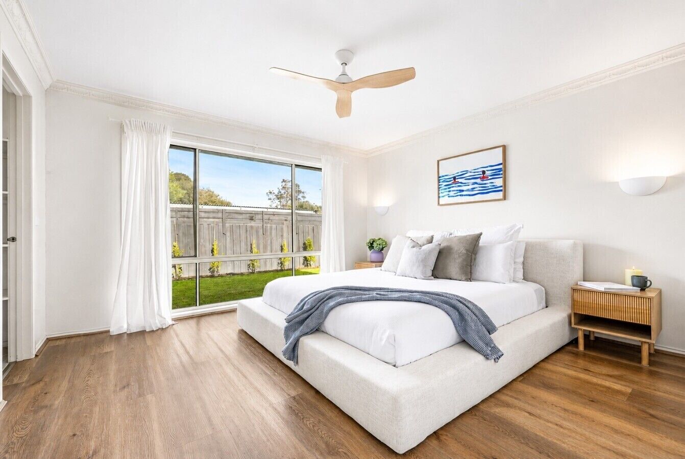 A bedroom with a bed, a ceiling fan, a nightstand, and a view of a garden through a sliding door.