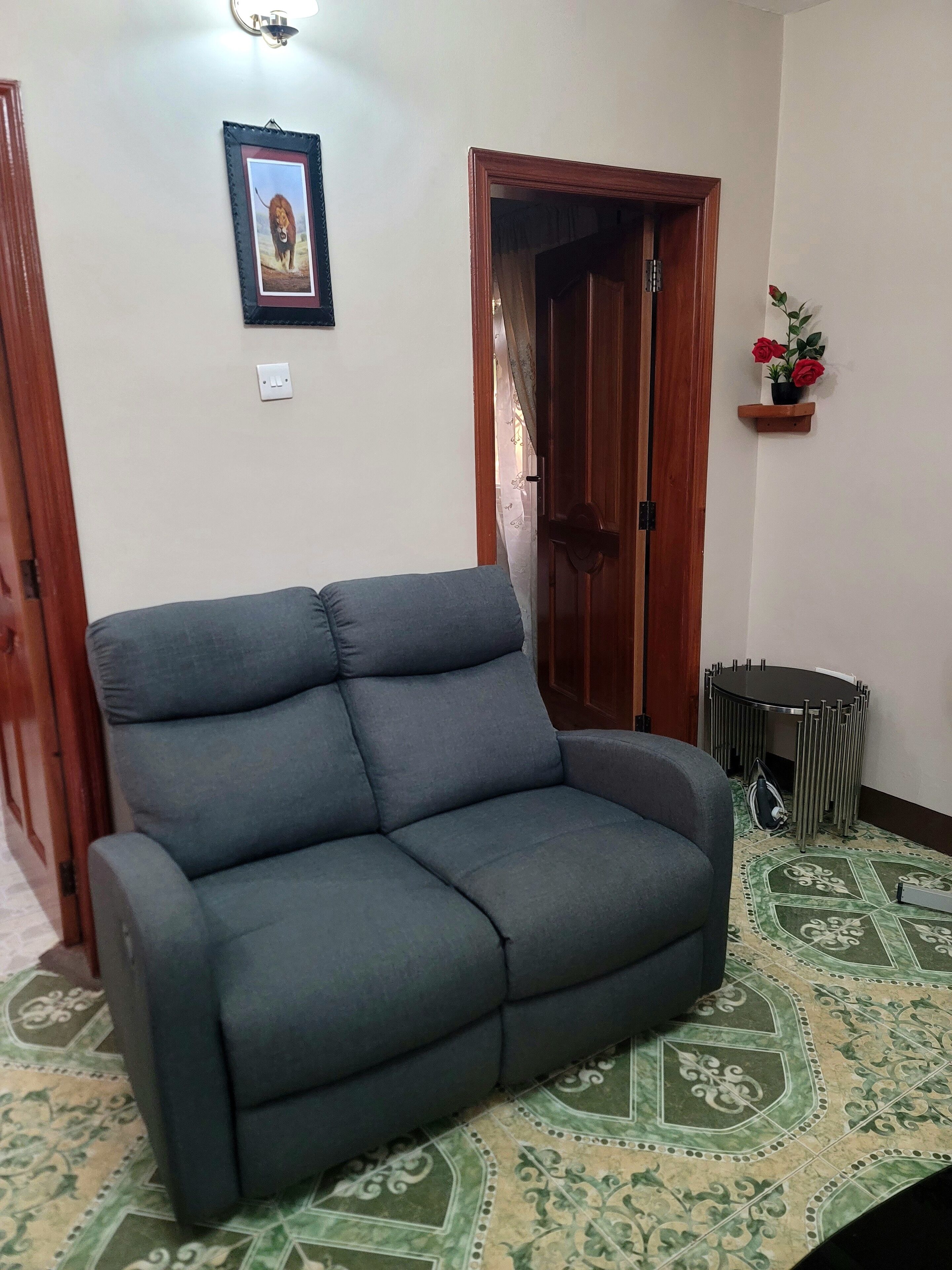 A living room with a grey sofa, a wooden door, a small wooden shelf with red flowers, and patterned floor tiles.