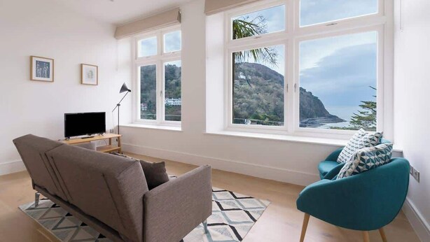A living room with a sofa, a chair, a TV, and large windows offering a view of a coastal landscape.