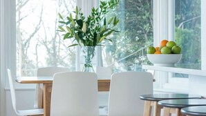 A dining area with a table and chairs, a vase with green foliage, and a bowl of fruit on a windowsill.